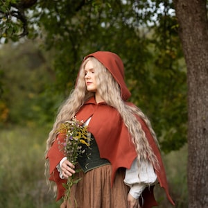 May include: A rust-colored hooded cape with a scalloped edge, paired with a green corset and a brown skirt. The person is holding a bouquet of wildflowers. The outfit has a medieval or fantasy theme.