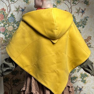 May include: A bright yellow hooded cape featuring a leaf-like design with stitched veins radiating from the center. The garment has a pointed hem and a structured hood, worn over a long-sleeved olive green shirt and a tiered brown skirt.