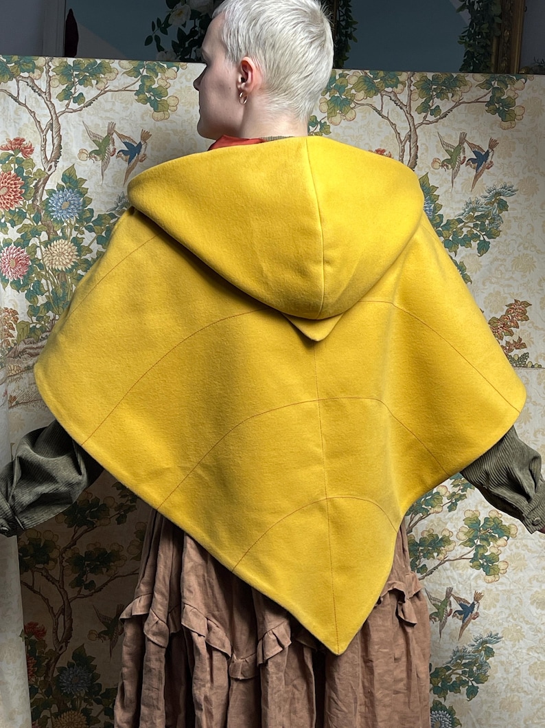 May include: A bright yellow hooded cape featuring a leaf-like design with stitched veins radiating from the center. The garment has a pointed hem and a structured hood, worn over a long-sleeved olive green shirt and a tiered brown skirt.