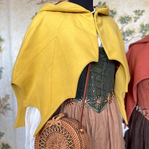 May include: A mustard yellow hooded cape with leaf-shaped edges, paired with a green corset, brown skirt, and a round woven handbag. The cape is the focal point, with detailed stitching. The outfit is displayed on a mannequin.