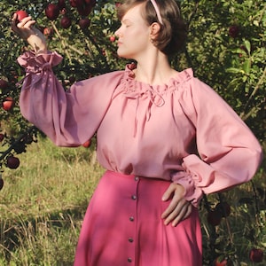 May include: A woman wearing a pink blouse with a ruffled neckline and long sleeves, paired with a pink skirt with a button-up front. She is standing in an orchard with apple trees.