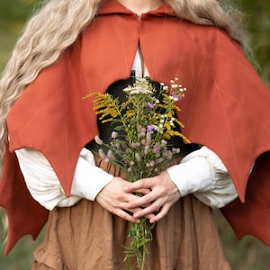 May include: A rust-colored hooded cape with leaf-shaped edges. The cape is worn over a white blouse and a brown skirt. A bouquet of wildflowers is held in front of the cape.