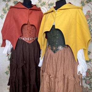 May include: Two medieval-style outfits on display. One features a rust-colored hooded cape, a brown corset, and a tiered brown skirt. The other outfit has a yellow cape, a green corset, and a tiered brown skirt. Both outfits have white blouses.