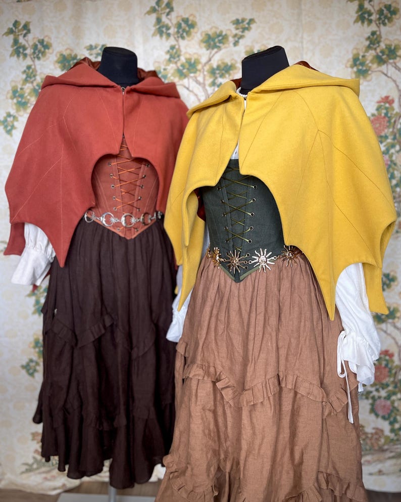 May include: Two medieval-style outfits on display. One features a rust-colored hooded cape, a brown corset, and a tiered brown skirt. The other outfit has a yellow cape, a green corset, and a tiered brown skirt. Both outfits have white blouses.