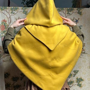 May include: A mustard yellow hooded cape featuring a pointed hemline and curved seam detailing across the back. The garment has a large, structured hood and a triangular shape that drapes over the shoulders, crafted from a solid, smooth fabric.