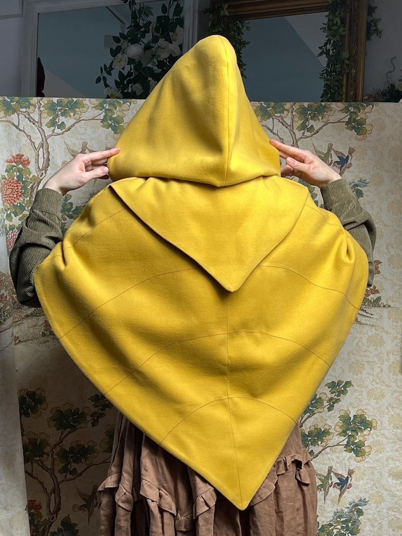 May include: A mustard yellow hooded cape featuring a pointed hemline and curved seam detailing across the back. The garment has a large, structured hood and a triangular shape that drapes over the shoulders, crafted from a solid, smooth fabric.