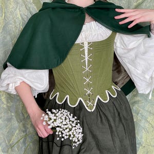 May include: A dark green hooded cape worn over an olive green corset with white lacing and a scalloped hem. Underneath is a white blouse with voluminous sleeves and a dark green skirt. A small bunch of white baby's breath flowers is held in one hand.
