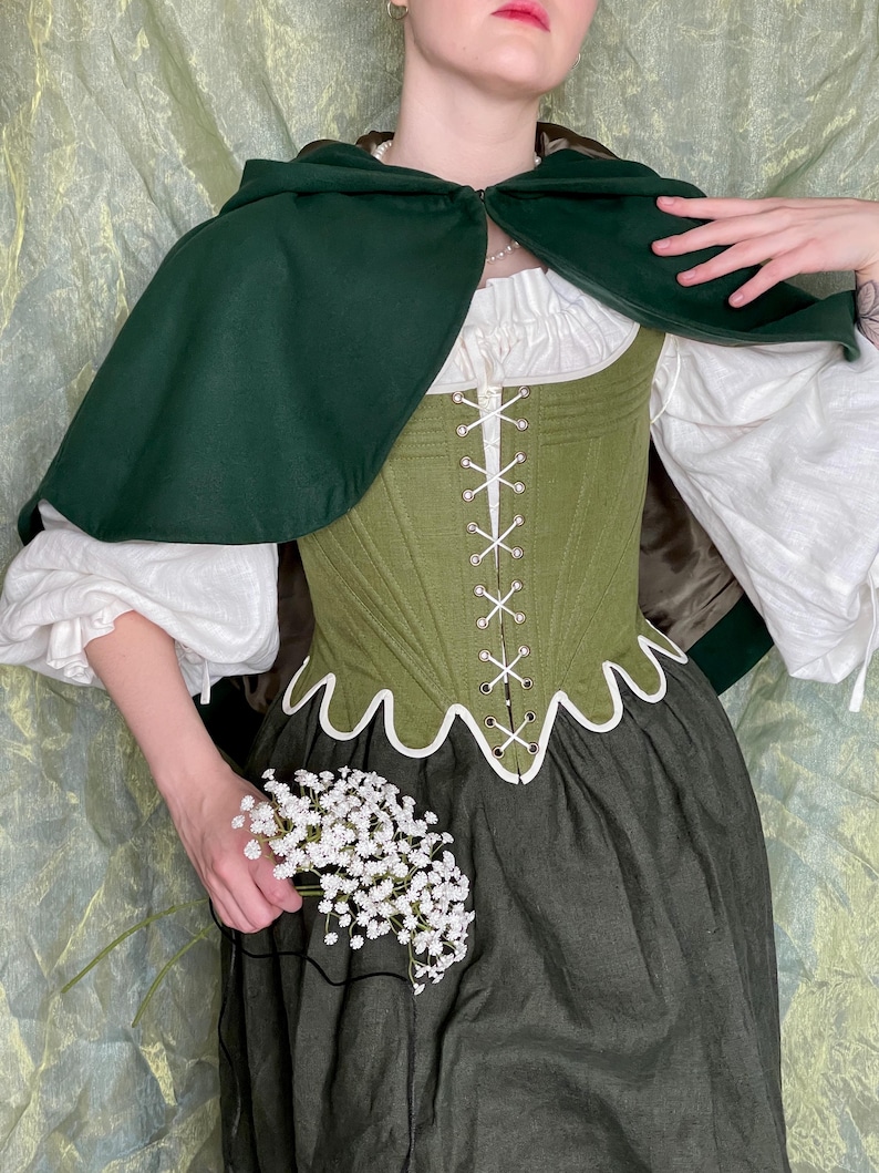 May include: A dark green hooded cape worn over an olive green corset with white lacing and a scalloped hem. Underneath is a white blouse with voluminous sleeves and a dark green skirt. A small bunch of white baby's breath flowers is held in one hand.
