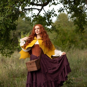 May include: A woman in a mustard yellow cape, a brown corset, and a long, ruffled plum skirt stands in a field. She carries a wooden box. Her red hair cascades down her shoulders. The scene is set outdoors with a tree overhead.