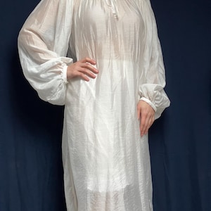 May include: A white, semi-sheer, knee-length dress with long, voluminous bishop sleeves gathered at the wrists. The garment features a round neckline with a central tie closure and a loose, flowing silhouette made of a lightweight, slightly textured fabric.