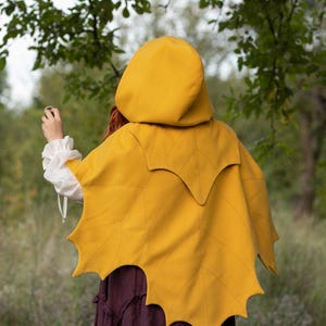 May include: A mustard yellow hooded cape with a leaf-shaped design on the back. The cape is made of a soft fabric and is worn over a white blouse and a dark purple skirt. The background features green foliage.