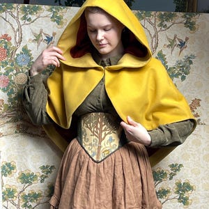 May include: A mustard yellow hooded cape with a deep red interior lining, worn over an olive green corduroy shirt and a corset featuring a tree and floral pattern, paired with a brown tiered skirt with a ruffled hemline detail.