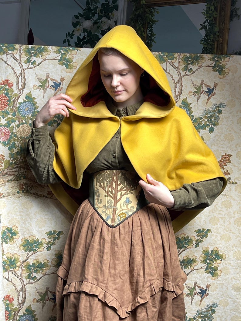 May include: A mustard yellow hooded cape with a deep red interior lining, worn over an olive green corduroy shirt and a corset featuring a tree and floral pattern, paired with a brown tiered skirt with a ruffled hemline detail.