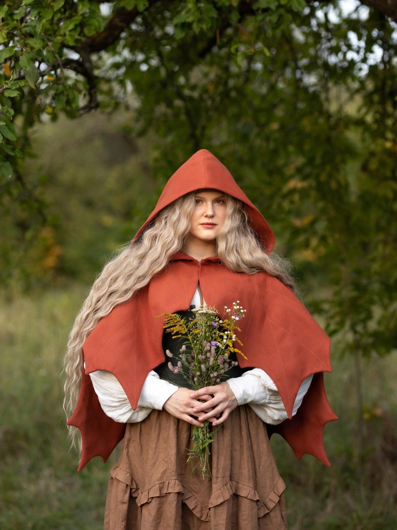 May include: A rust-colored hooded cape with pointed edges, worn over a white blouse and brown skirt. The cape is the focal point, with a woman holding a bouquet of wildflowers. The background is a blurred green forest.