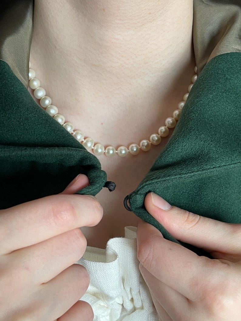 May include: A single strand of off-white, round pearls worn as a necklace. The pearls are uniform in size and strung together. The necklace is positioned around the neck, resting against the skin, visible above a dark green garment and white fabric.