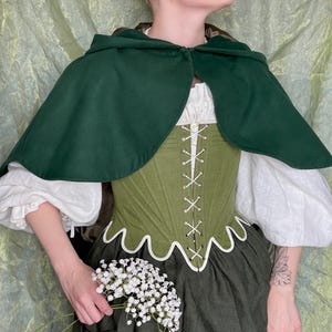 May include: A dark green hooded cape worn over a white long-sleeved blouse and an olive green corset with white lacing and a scalloped hem. The outfit includes a dark green skirt and is accented by a small bouquet of white baby's breath flowers.