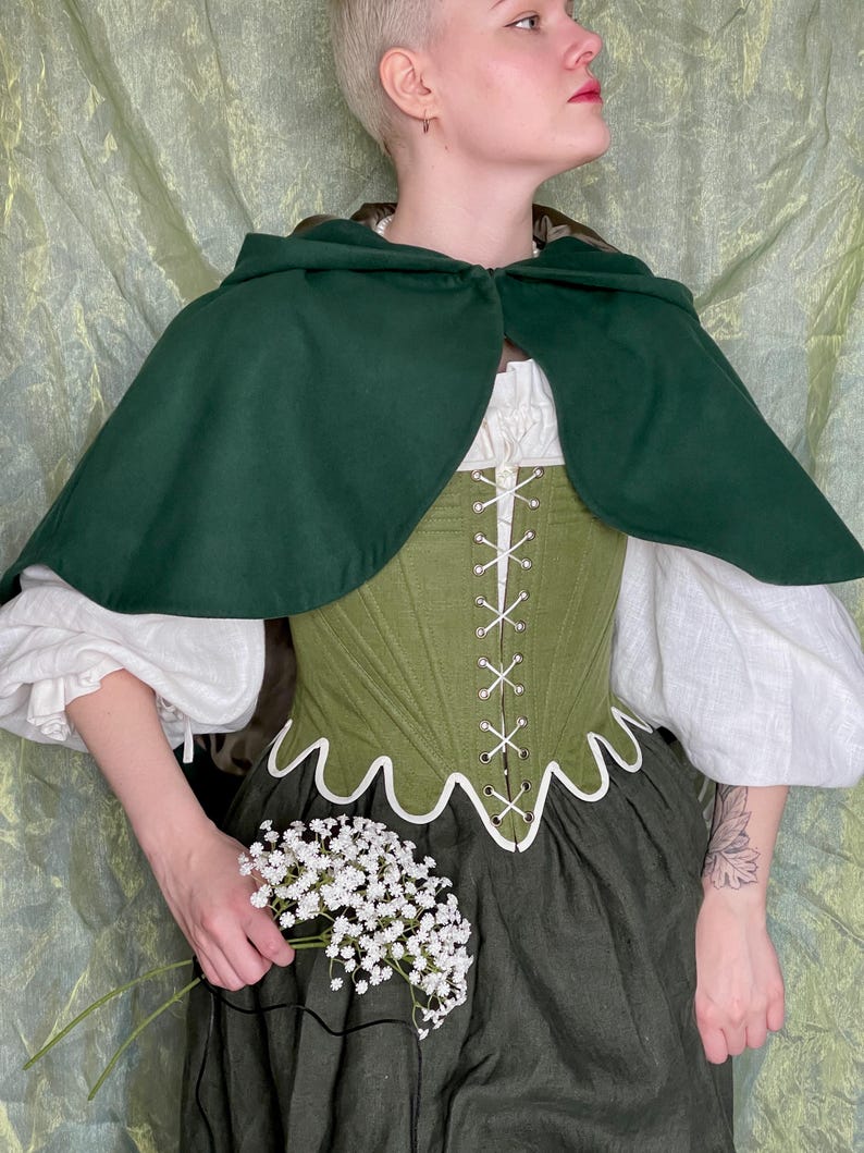May include: A dark green hooded cape worn over a white long-sleeved blouse and an olive green corset with white lacing and a scalloped hem. The outfit includes a dark green skirt and is accented by a small bouquet of white baby's breath flowers.