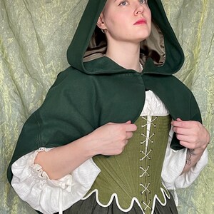 May include: A dark green hooded cape with a tan lining worn over a white long-sleeved blouse and an olive green corset with white scalloped trim and front lacing, paired with a dark gray skirt, creating a layered ensemble of various textures.