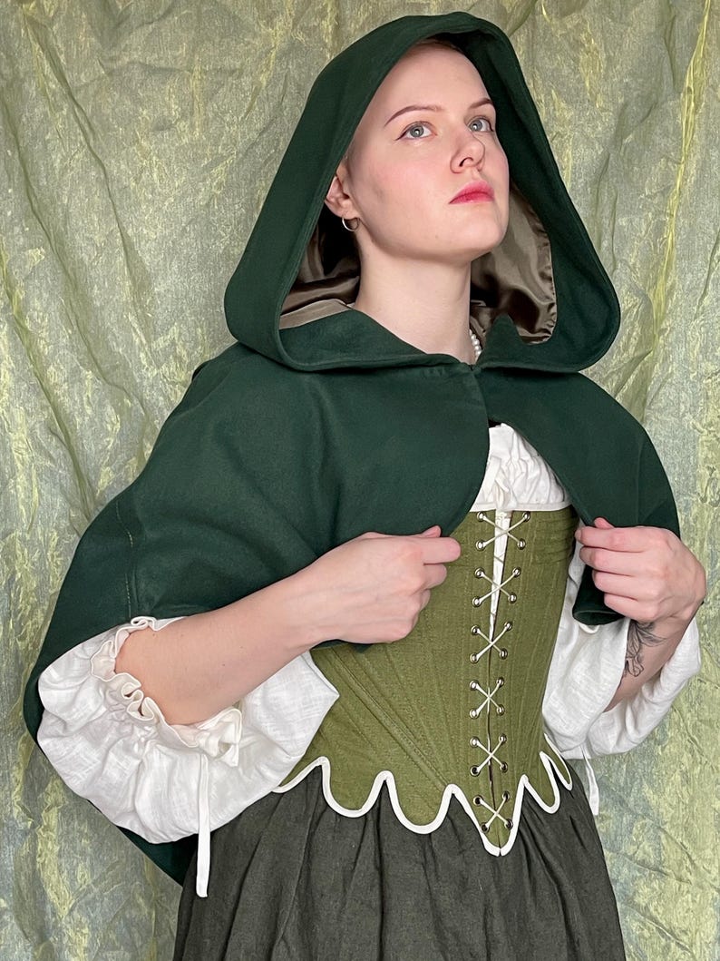 May include: A dark green hooded cape with a tan lining worn over a white long-sleeved blouse and an olive green corset with white scalloped trim and front lacing, paired with a dark gray skirt, creating a layered ensemble of various textures.