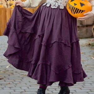 May include: A woman wearing a purple, tiered skirt with ruffles. She is holding a carved pumpkin with a smiling face. The skirt is long and flowing, and the pumpkin is orange with black details.