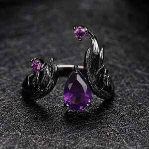 May include: A black ring with a large purple teardrop-shaped gemstone and two smaller purple gemstones. The ring features a detailed design resembling wings.