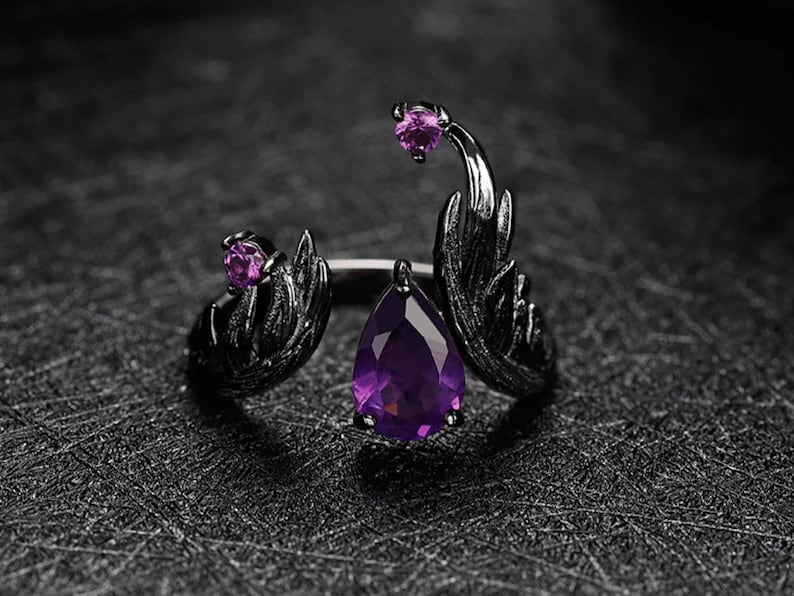 May include: A black ring with a large purple teardrop-shaped gemstone and two smaller purple gemstones. The ring features a detailed design resembling wings.