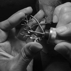 May include: A person's hands are holding a small, intricate metal tree sculpture. The person is using a rotary tool to refine the details of the sculpture.