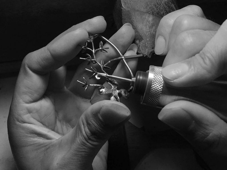 May include: A person's hands are holding a small, intricate metal tree sculpture. The person is using a rotary tool to refine the details of the sculpture.