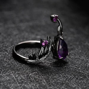 May include: Black ring with a purple gemstone and intricate leaf-like details. The ring has a delicate, organic design.