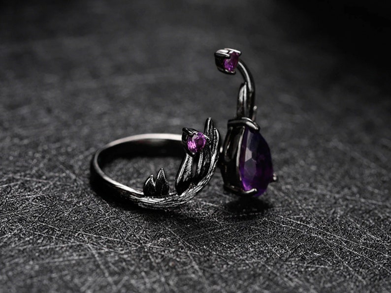 May include: Black ring with a purple gemstone and intricate leaf-like details. The ring has a delicate, organic design.
