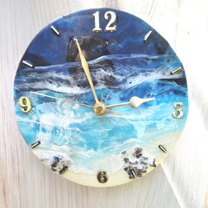 Op de afbeelding: Een ronde wandklok met een harskunstontwerp van een strandscène met diepblauw water, witte brekende golven en een zandstrand. De wijzerplaat heeft goudkleurige wijzers en goudkleurige cijfers op de posities 12, 3, 6 en 9.