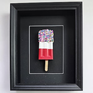 May include: A framed wall art piece featuring a three-dimensional popsicle sculpture. The popsicle has a red base, a white middle section, and a top covered in multicolored sprinkles, all attached to a wooden stick, set against a black background.