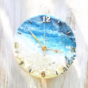 May include: A round wall clock featuring a resin art design of a beach scene with blue ocean waves, white sea foam, and a sandy shore. The clock face includes gold-colored numbers at the 12, 3, 6, and 9 positions, gold hands, and small decorative stones.