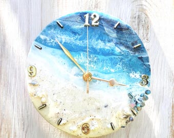 May include: A round wall clock featuring a resin art design of a beach with blue ocean waves, white sea foam, and a sandy shore. The clock face includes gold-coloured numbers at the 12, 3, 6, and 9 positions, gold hands, and small decorative stones.