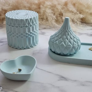May include: A set of three light blue decorative boxes with geometric patterns. The boxes are made of concrete and have a smooth, matte finish. The set includes a round box, a heart-shaped box, and a teardrop-shaped box. The boxes are arranged on a white marble surface.