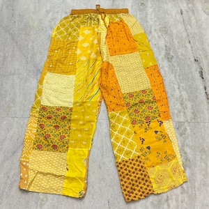 Op de afbeelding: Gele patchworkbroek met een tailleband met trekkoord. De broek heeft een verscheidenheid aan gele en oranje stoffen vierkanten met patronen, waaronder bloemen-, stippen- en geometrische ontwerpen. De broek ligt plat op een lichtgrijze ondergrond.