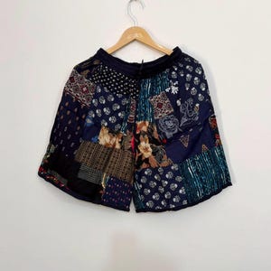 May include: A pair of dark blue shorts featuring a patchwork design with various patterns including floral, geometric, and polka dot prints. The shorts have an elastic waistband with a drawstring and are displayed on a wooden clothes hanger.