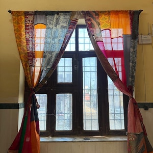 Puede incluir: Una ventana enmarcada en madera oscura, adornada con cortinas de patchwork. Las cortinas son una mezcla de telas naranjas, amarillas, azules y rojas. Las cortinas están atadas, revelando la ventana y la luz.