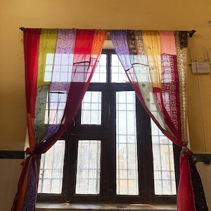 May include: A colorful patchwork curtain hangs in front of a dark wooden window. The curtain is made of various fabrics in red, orange, yellow, green, and purple. The curtain is tied back on both sides.