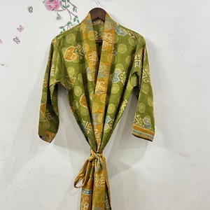 May include: A green and yellow patterned robe hangs on a wooden hanger. The robe has a floral and geometric design in shades of green, yellow, and blue. The robe is tied at the waist with a matching sash.