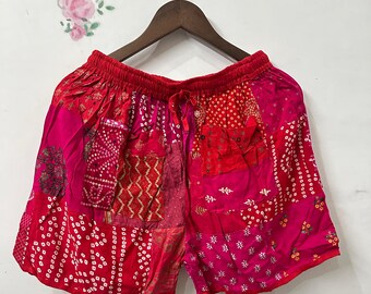 May include: Red and pink patchwork shorts with an elastic waistband and drawstring. The shorts feature various patterns and textures, including dotted and geometric designs. The garment is hanging on a wooden hanger.