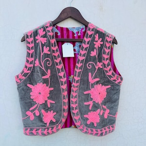 May include: A gray velvet vest with bright pink floral embroidery. The vest is reversible, with a striped pink and red interior and a patterned blue and white fabric. The vest is hanging on a wooden hanger.