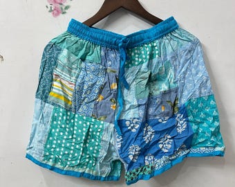 May include: Patchwork shorts in shades of blue, teal, and white, with a drawstring waist. The shorts are made from various fabric patterns, creating a unique, colorful design. The shorts are hanging on a wooden hanger.