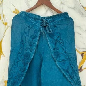 May include: Teal blue wrap pants featuring a wide-leg silhouette and an overlapping front panel. The garment is decorated with dark teal floral and vine embroidery along the edges and includes a drawstring tie closure at the center waist.