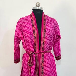 Pode incluir: Um roupão magenta de manga comprida e comprimento até o joelho com um padrão geométrico repetitivo num tom de rosa mais claro. A peça inclui um cinto de amarrar a condizer e acabamentos decorativos ao longo da abertura frontal.