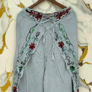 May include: Light gray wrap pants featuring a decorative waist tie and intricate floral embroidery in red, green, and black thread along the edges. The garment has a split-leg design with scalloped trim and is displayed on a wooden hanger.