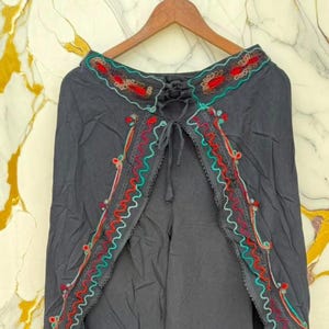 May include: Dark gray wrap-style pants featuring an open front design with a drawstring waist. The edges are decorated with intricate embroidery in red, teal, and light green, showcasing a wavy pattern and small circular accents throughout.