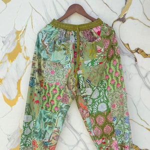 Puede incluir: Pantalones patchwork verdes con estampado floral y botánico. Los pantalones tienen cintura elástica y cordón. La tela presenta varios diseños en tonos verdes, rosas y azules. Colgados de una percha de madera.