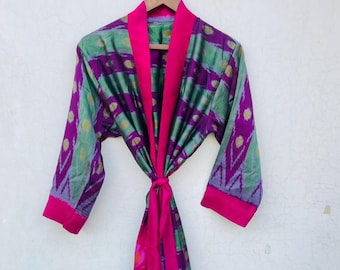 May include: A colourful patterned robe in green, purple, and pink, hanging on a wooden hanger. The robe has a repeating geometric design with a contrasting pink trim and tie closure. The fabric appears silky.
