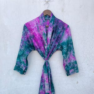 Könnte beinhalten: Ein Batik-Kimono in Lila-, Grün- und Weißtönen hängt an einem Holzbügel. Der Kimono ist aus leichtem Stoff gefertigt und hat lange Ärmel und einen Bindverschluss in der Taille. Das Batikmuster ist lebendig und auffällig.